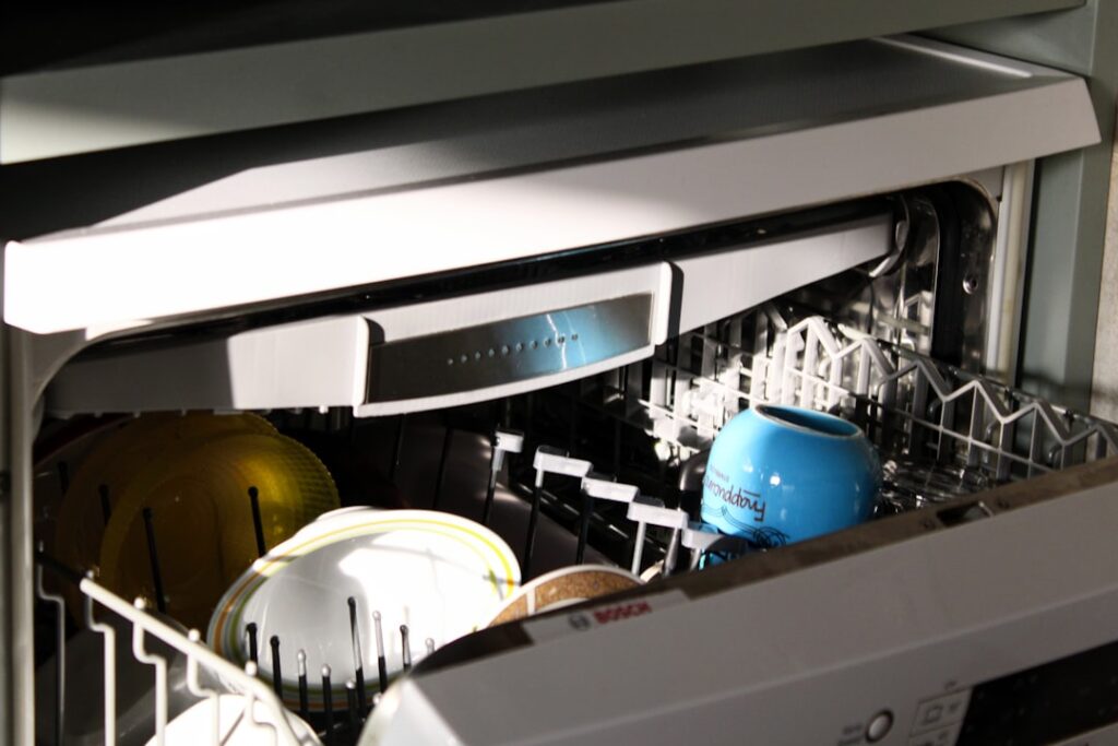 A person inspecting a built-in dishwasher in a residential kitchen, with the appliance door open and tools on the floor.